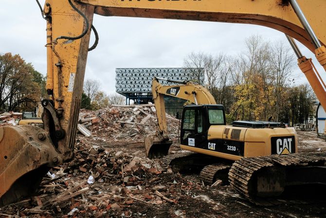 Two yellow construction excavators work on a debris-filled demolition site in front of a modern, patterned building.