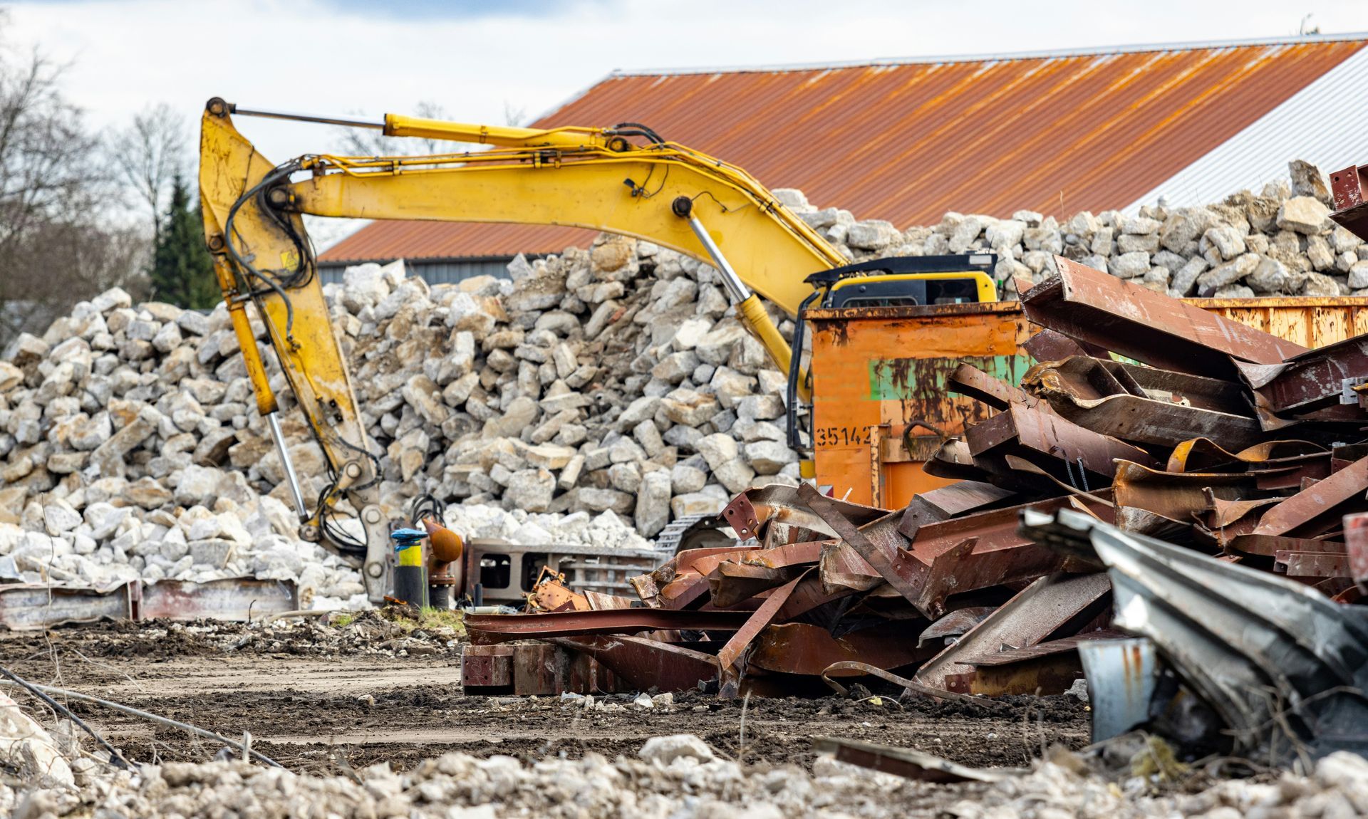 A yellow excavator sits in a construction yard filled with large piles of gray concrete rubble and rusted metal scrap.