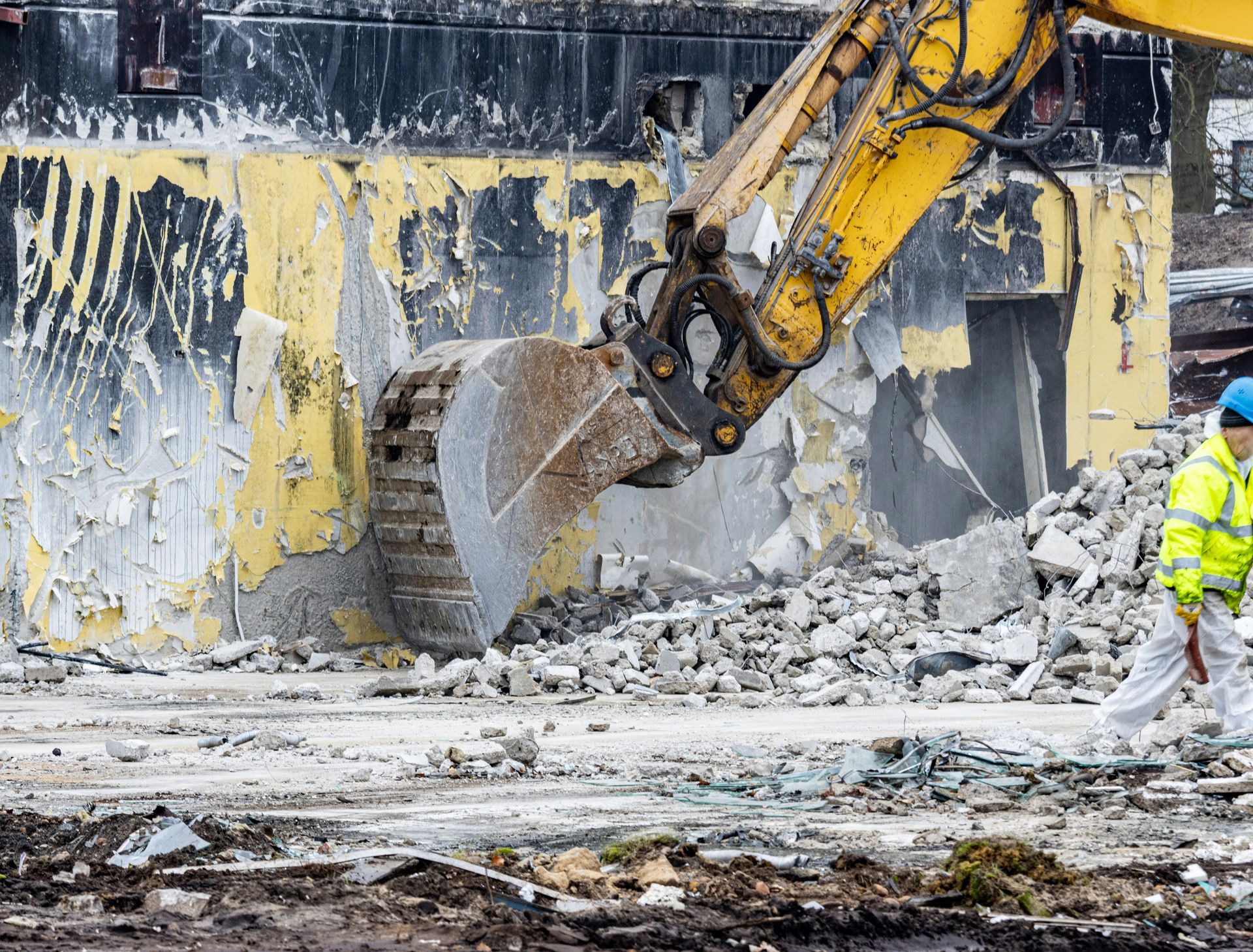 A yellow excavator demolishing a yellow-walled building, with a worker in a high-visibility jacket standing nearby.