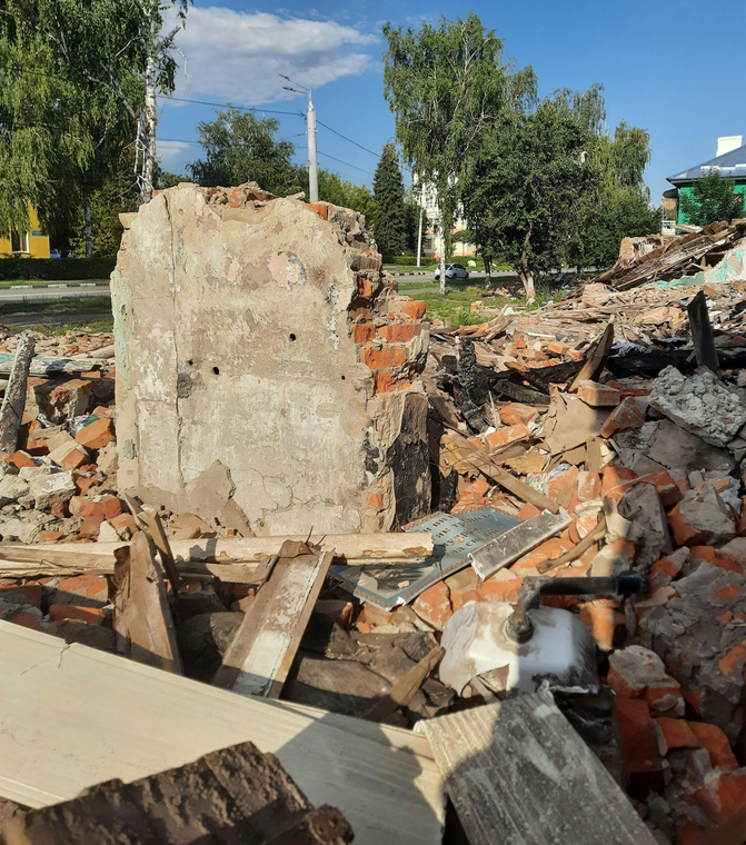 A large pile of rubble and broken brick walls from a demolished building, with trees and a clear blue sky in the background.