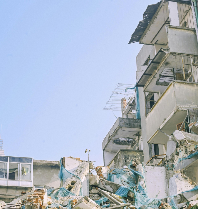 A partially collapsed multi-story concrete building with debris scattered in the foreground under a clear sky.