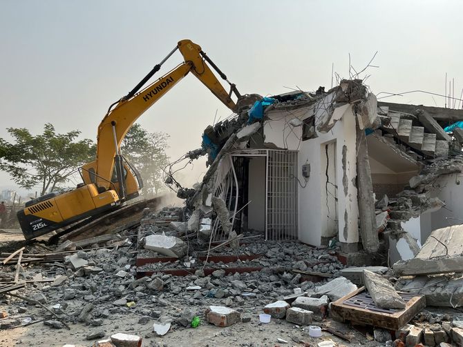 A yellow excavator demolishing a concrete building, surrounded by piles of rubble on a dusty, open site.