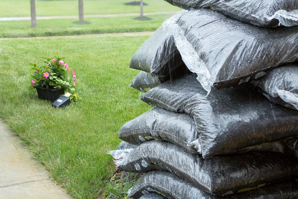 Stacked bags of mulch covered in plastic beside a lawn in the rain.