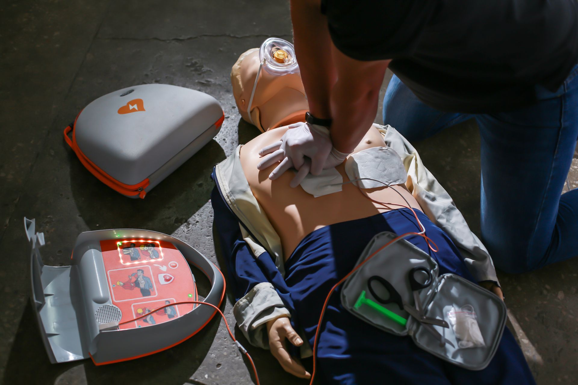Person performing CPR on a manikin beside an AED device and training pads outdoors