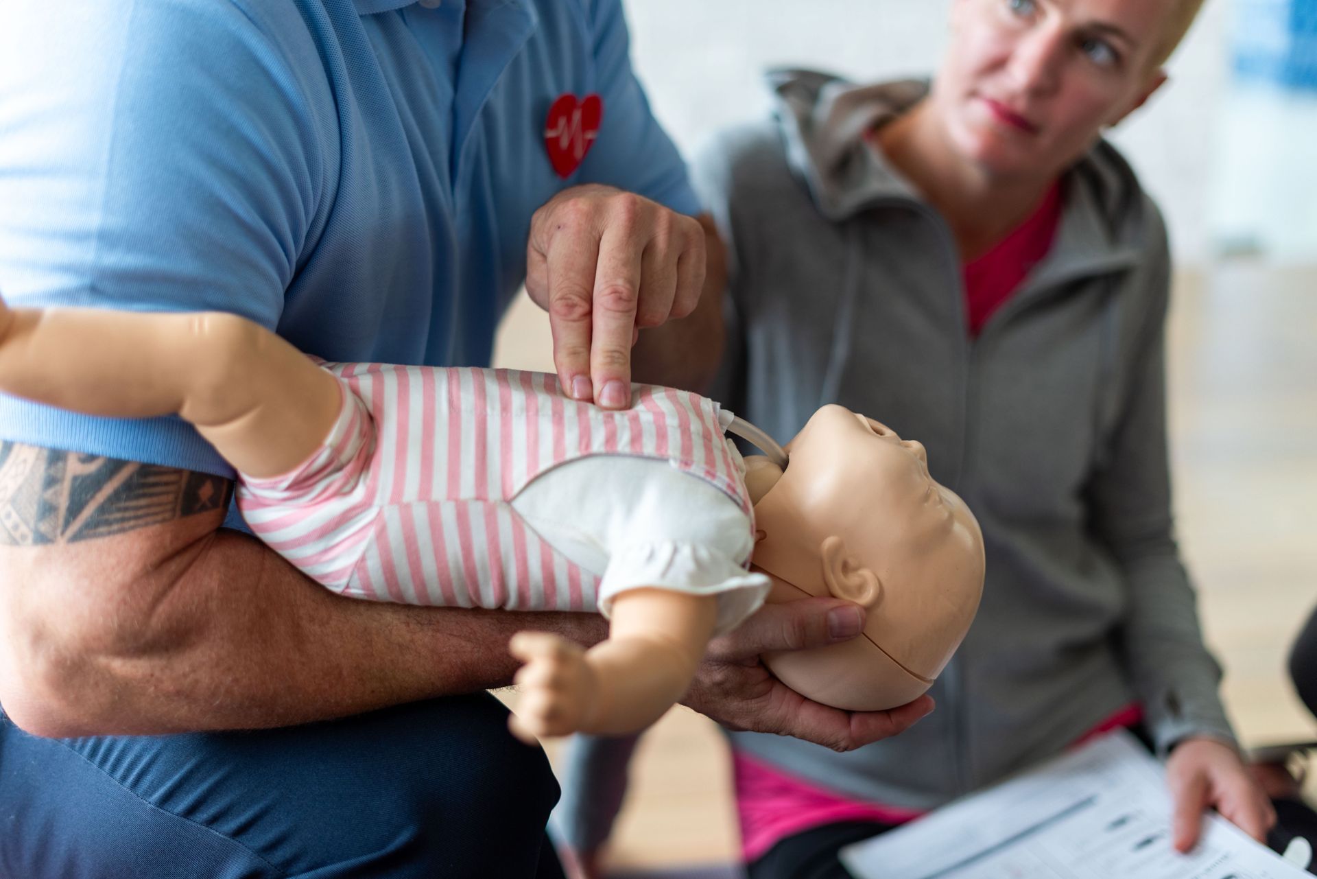 Caregiver demonstrating infant CPR on a baby doll while a woman observes and takes notes