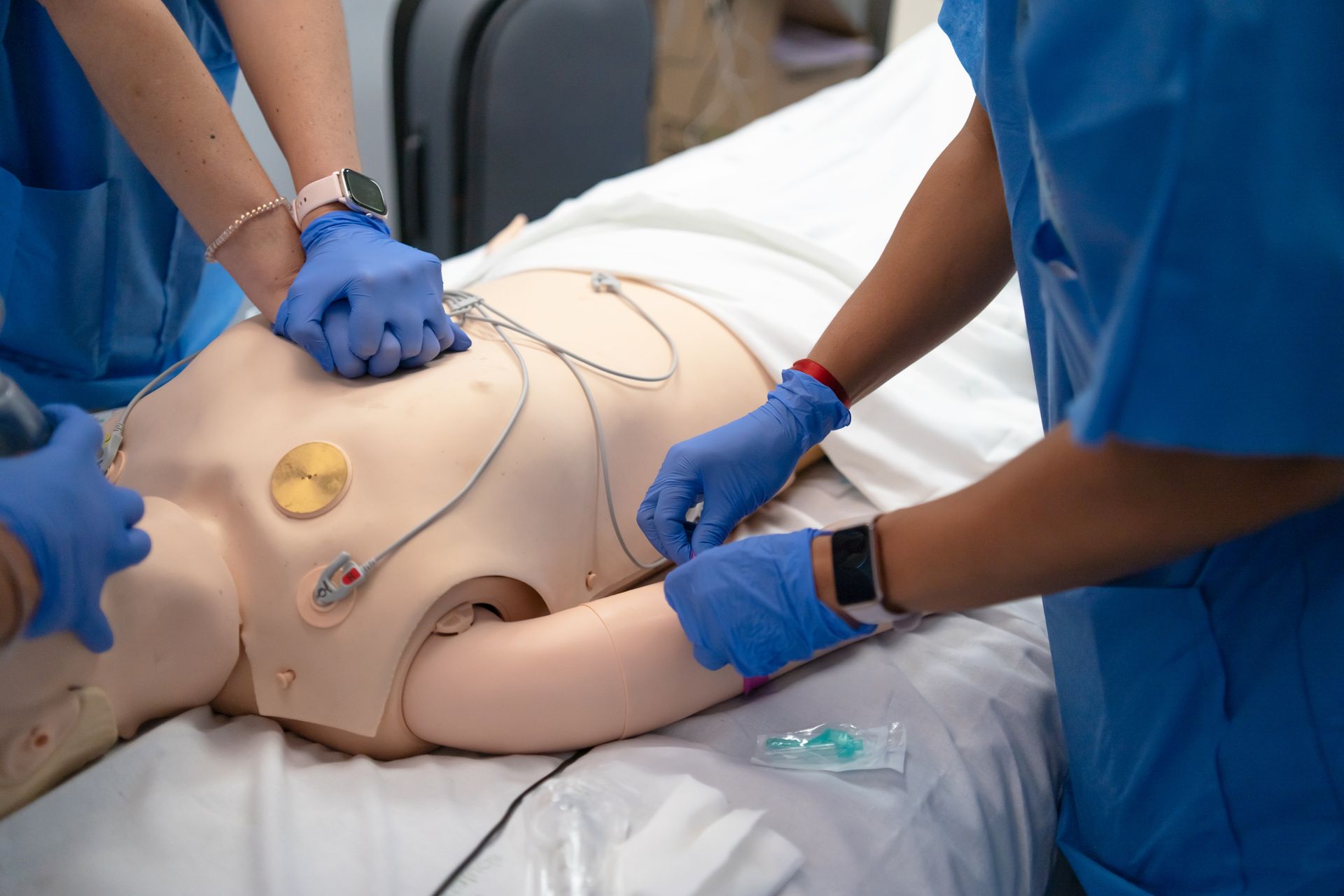 Medical training mannequin on a hospital bed with staff performing a procedure using defibrillator pads