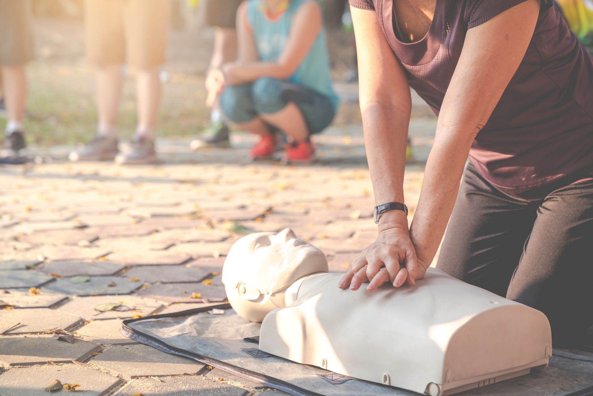 Person performing CPR on a mannequin outdoors while bystanders watch in the background