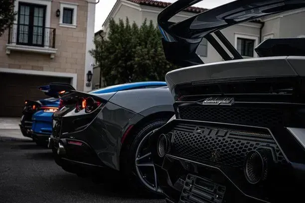 Three sports cars parked in front of a house: a blue, gray, and black model, showing rear detail.
