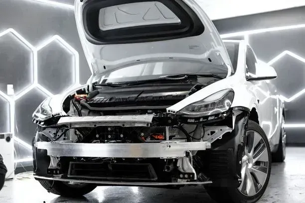 White Tesla Model Y with open hood and trunk, undergoing repair in a garage with honeycomb-shaped lights.