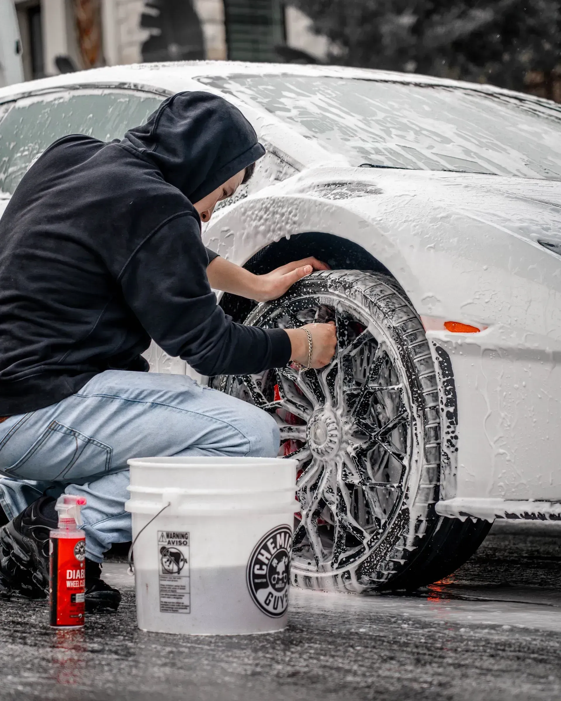 Man washing a white car's tire with soap, using a bucket, in an outdoor setting.