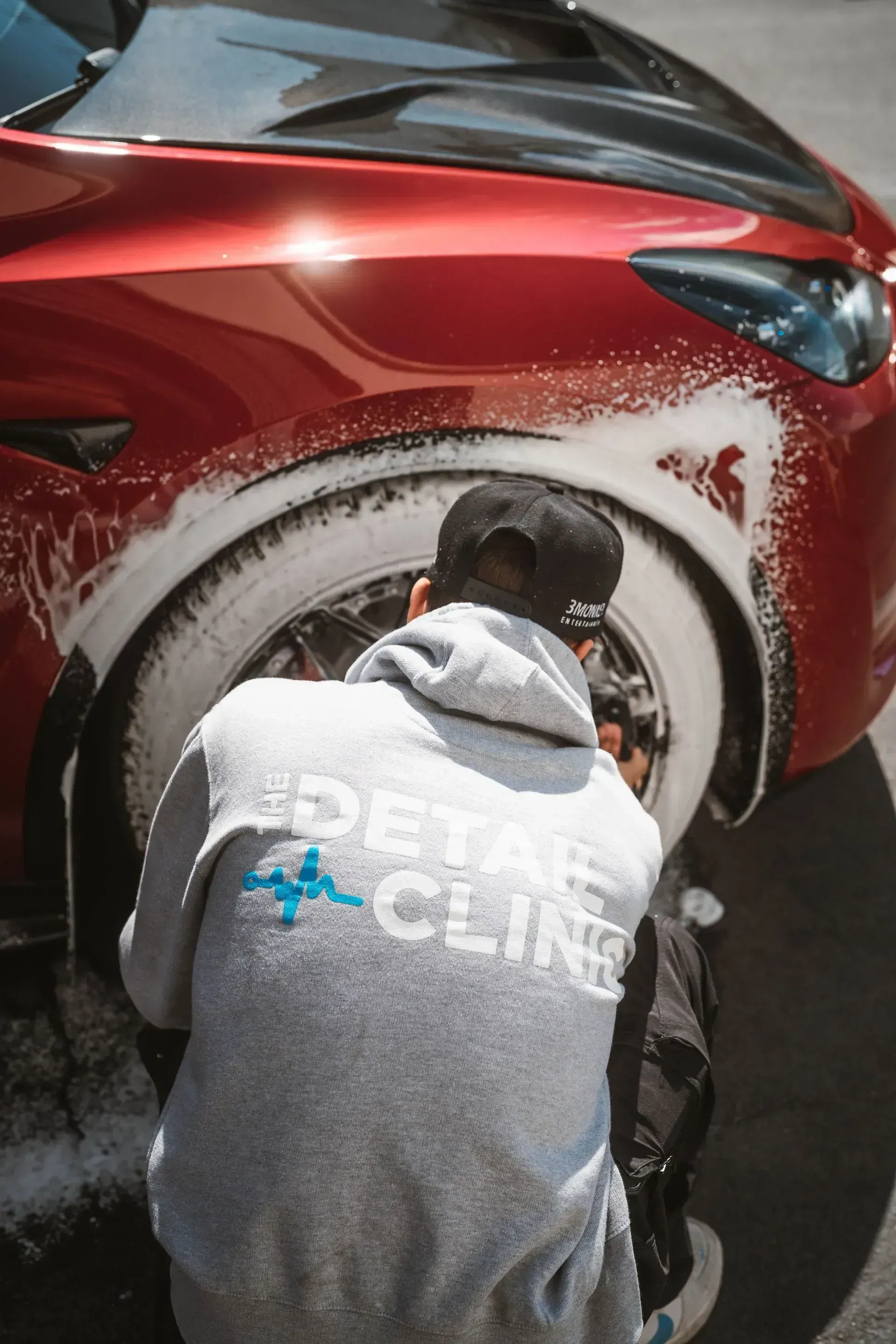 Person washing a red car with white foam. The person wears a grey hoodie with