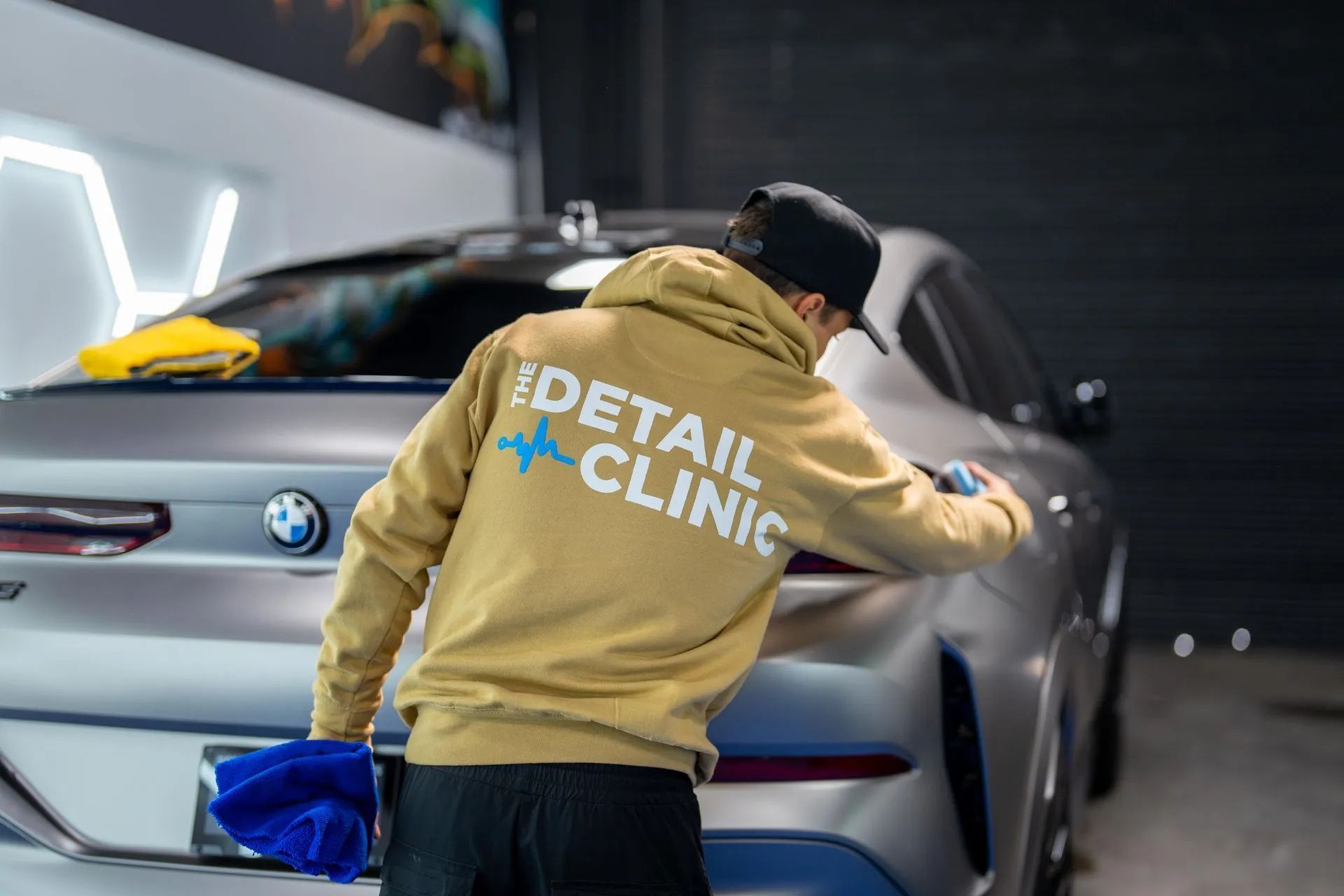 Person in tan hoodie cleaning a gray BMW car at the iDetail Clinic.