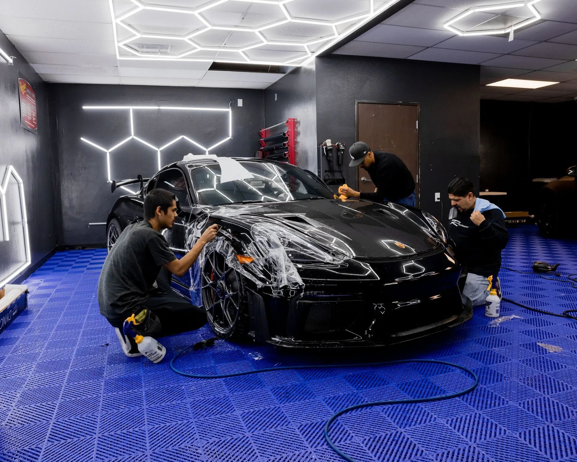 Three people applying film to a black car in a garage with blue floor tiles and hexagon lights.