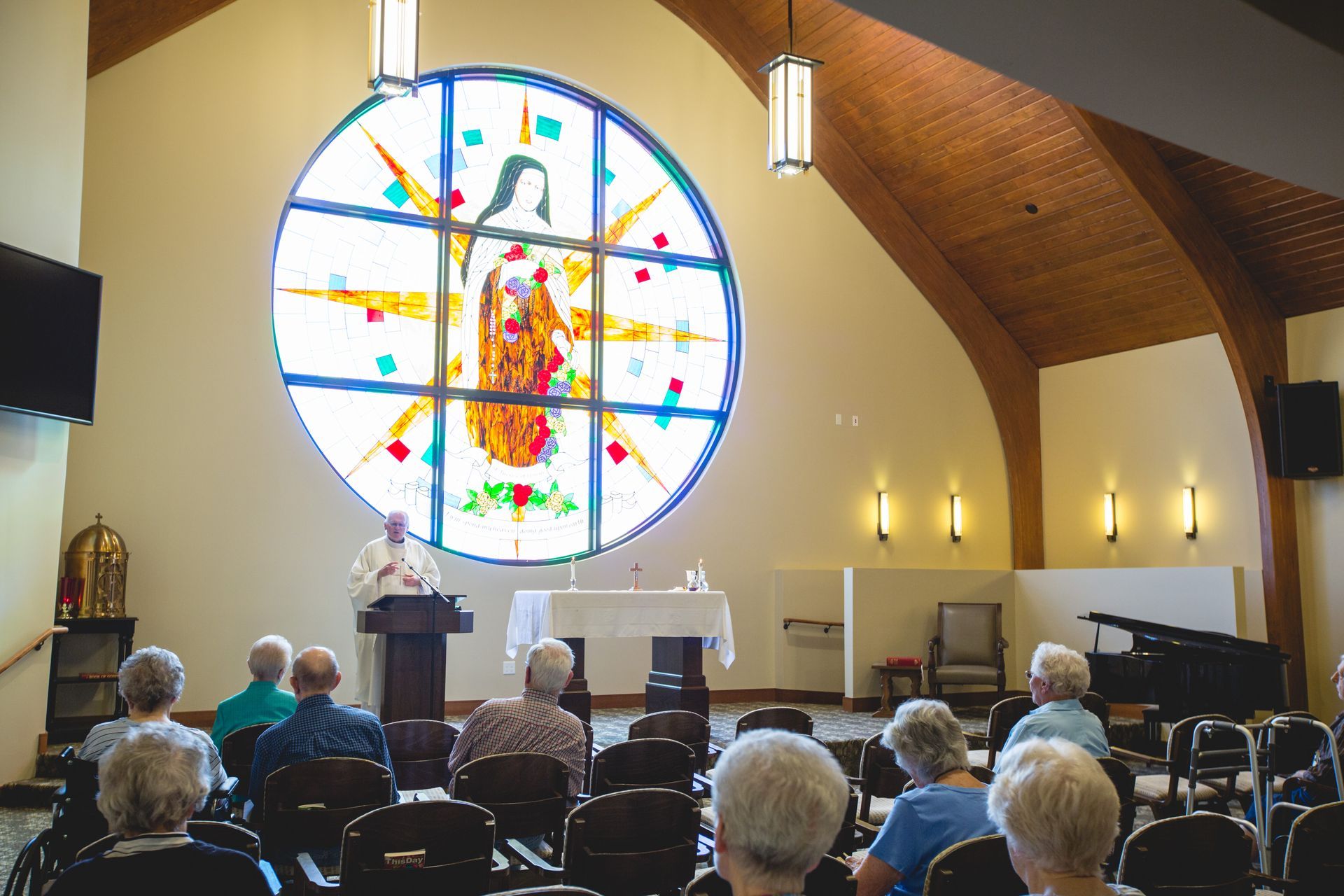 A priest leads a church service before a congregation, facing a large, illuminated stained-glass window of the Virgin Mary.