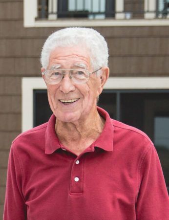Smiling person with short white hair and glasses, wearing a maroon polo shirt, standing outside in front of a building.