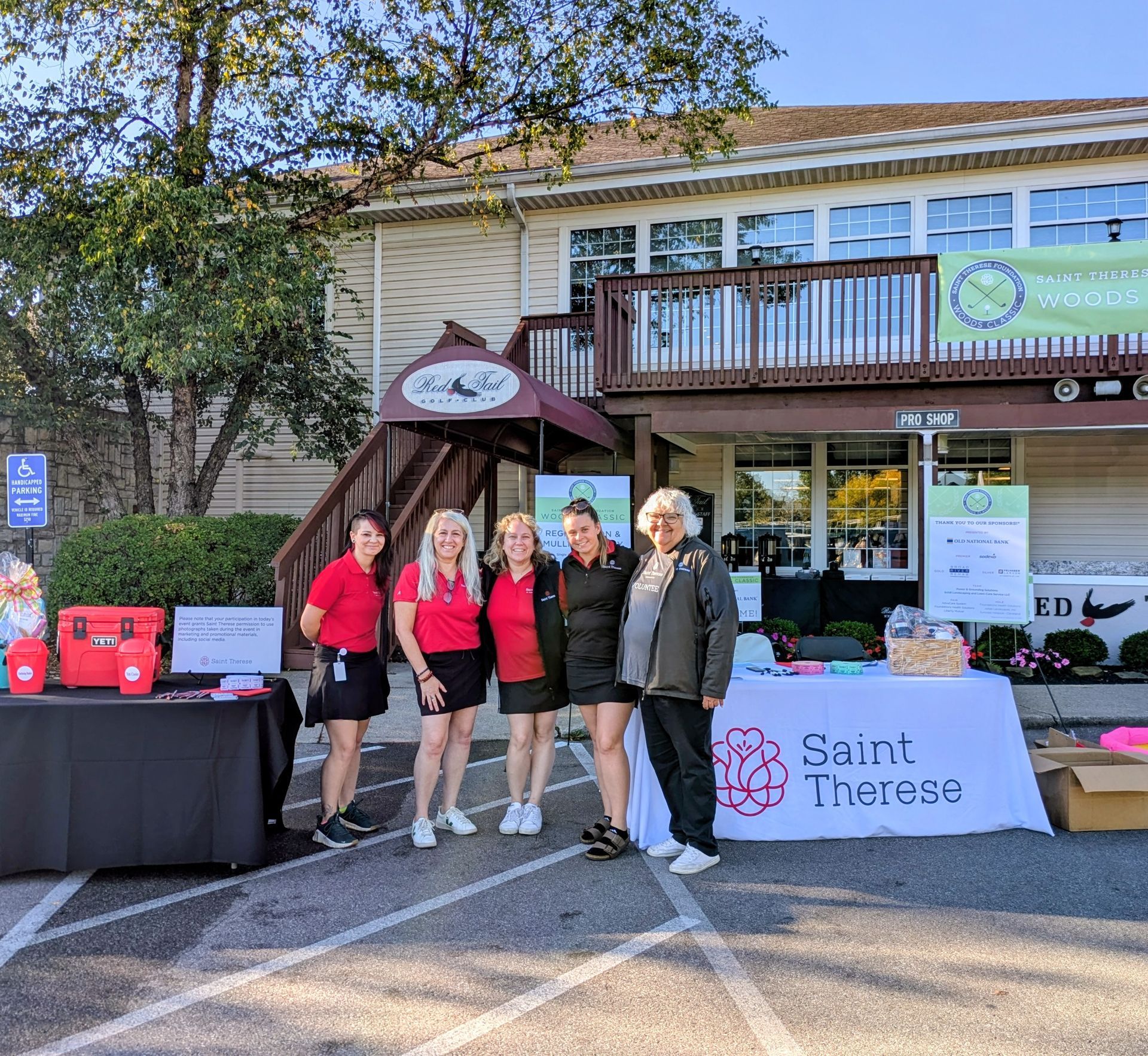 Five people stand in front of a Saint Therese event booth with a banner in an outdoor parking lot.