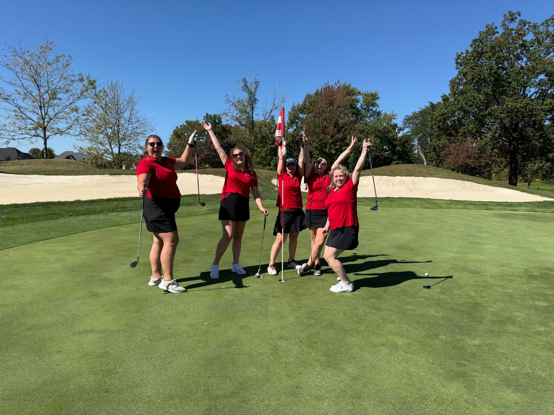 Four people in matching red shirts and black skirts pose excitedly around a golf flag on a sunny green.