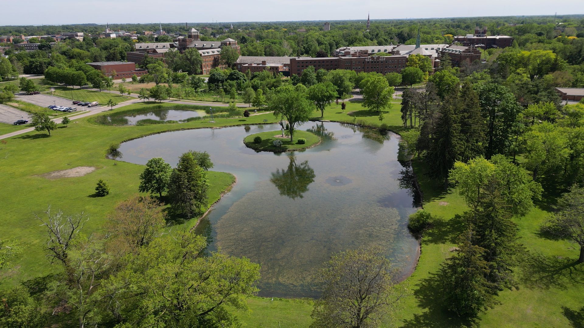 Aerial view of a green park featuring a pond with a small island, surrounded by trees and distant buildings.