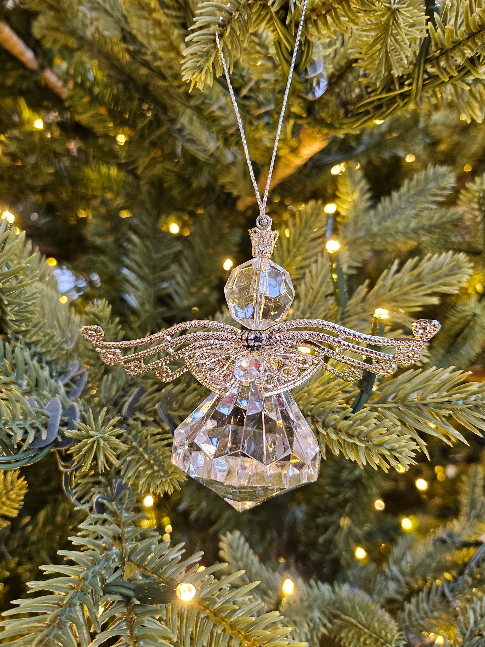 A clear crystal angel ornament with filigree gold wings hangs from a string on a lit Christmas tree.
