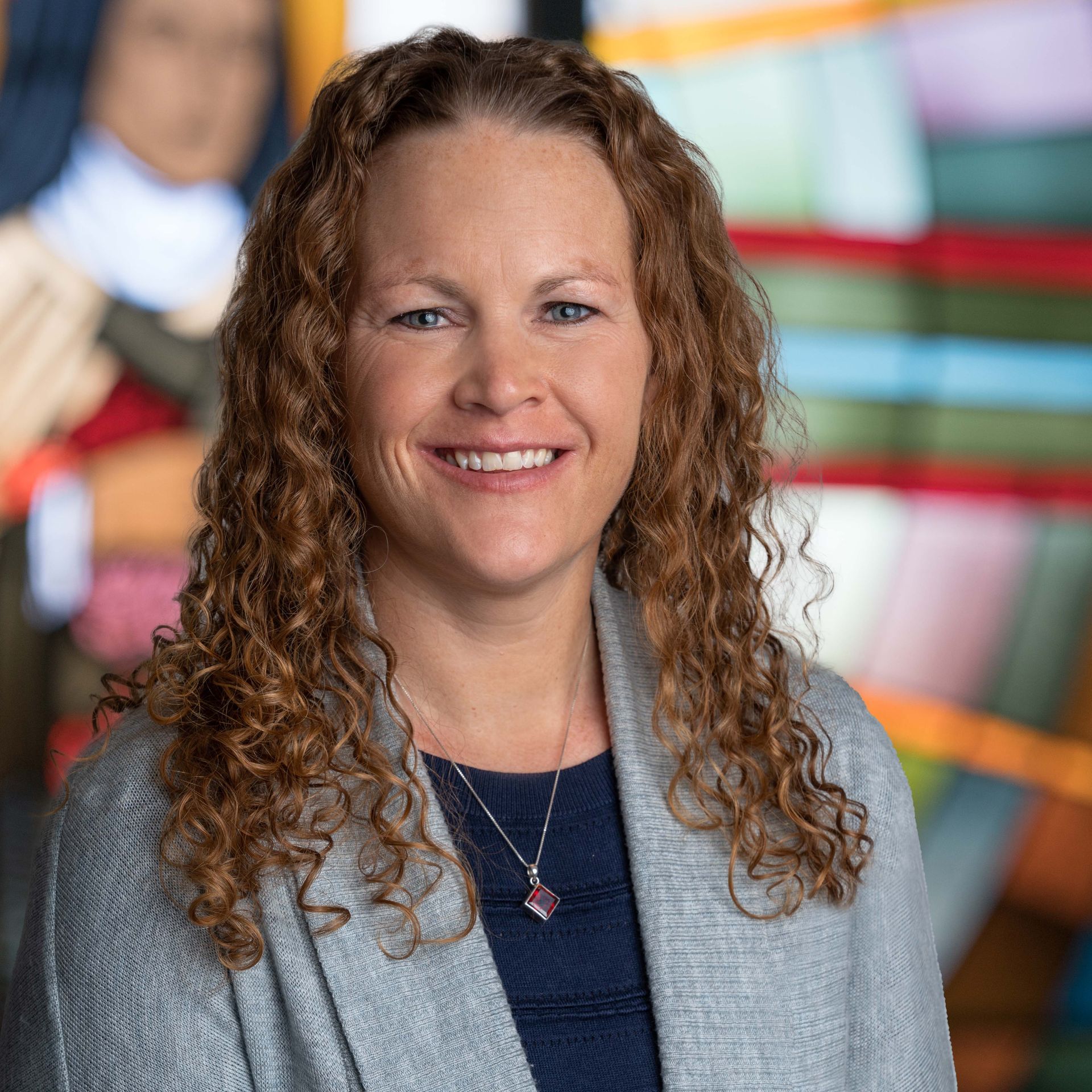 A person with curly hair smiles while wearing a grey cardigan and a necklace, set against a blurred, colorful background.