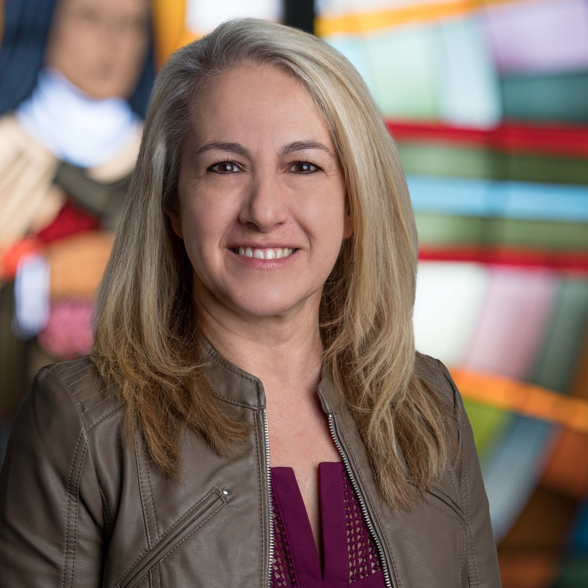 A smiling woman with shoulder-length blonde hair wearing a grey jacket over a maroon top, posed before a blurred backdrop.