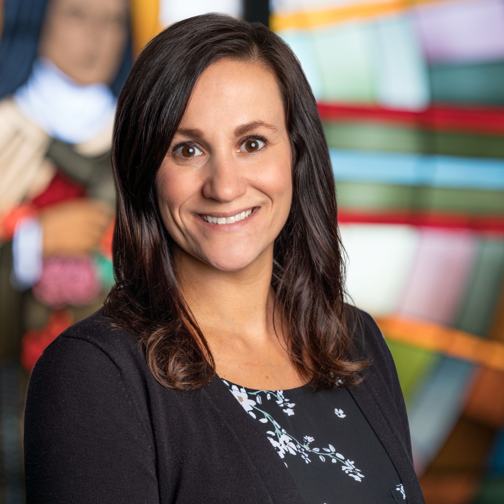 A person with dark hair smiling in a dark blazer over a patterned top, with a colorful, blurred background.
