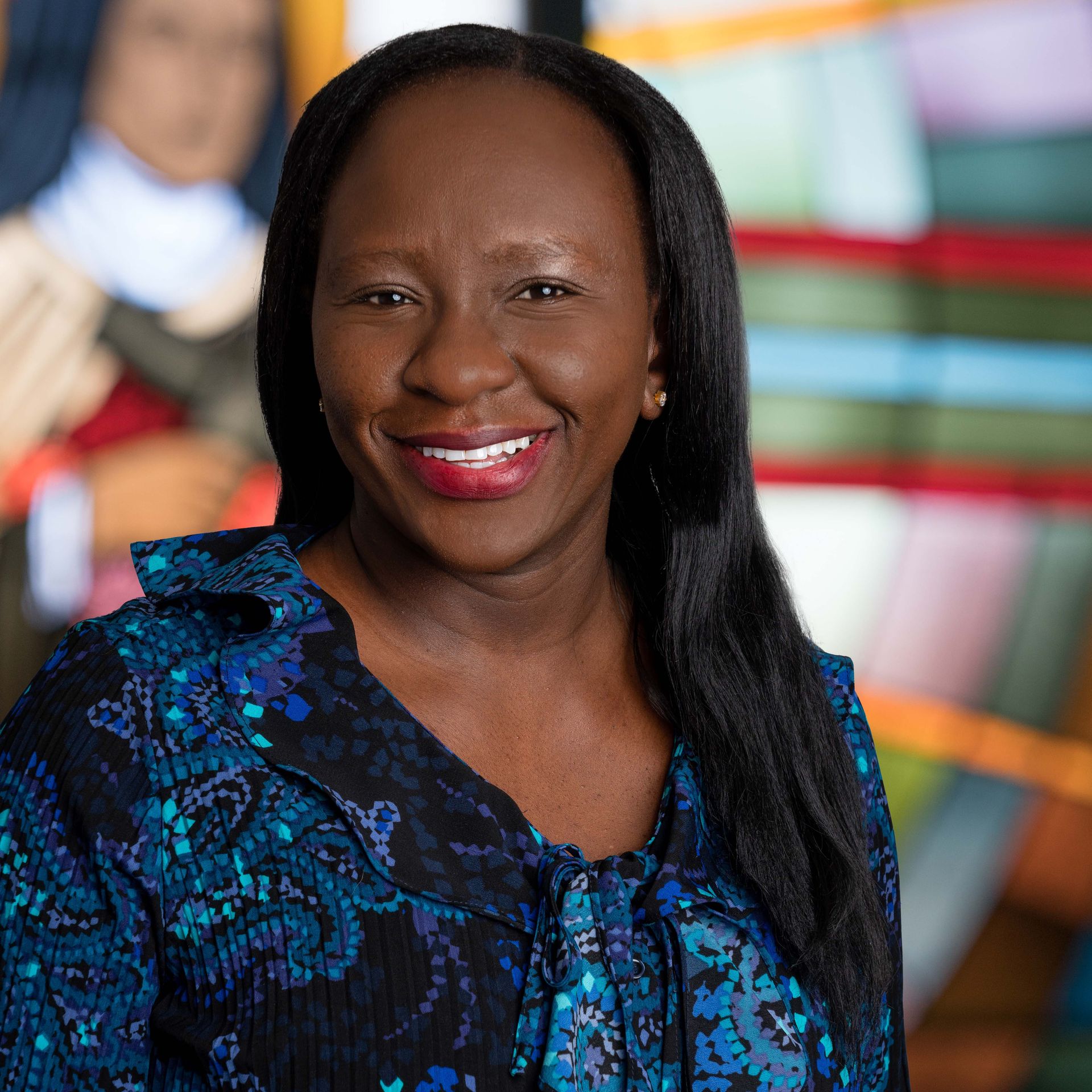 A smiling person wearing a blue patterned top, posed in front of a colorful, abstract background.