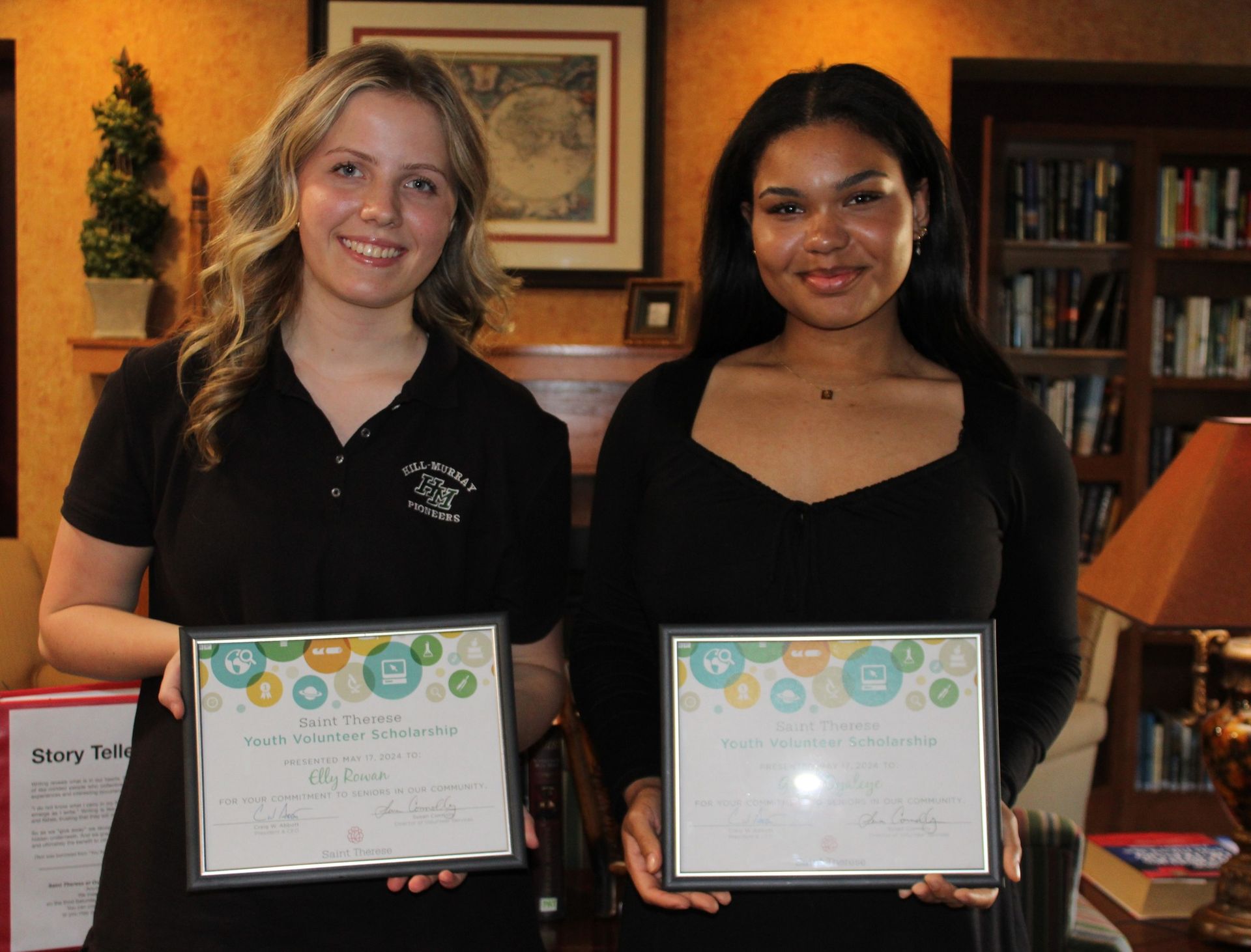 Two smiling people stand in a library, each holding a framed award certificate.