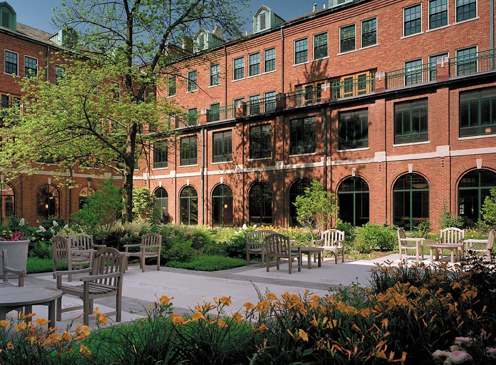 A red brick building with arched ground-floor windows overlooks a stone patio with outdoor seating and lush gardens.