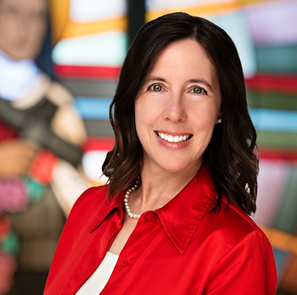 A smiling woman wears a red button-down shirt and a pearl necklace, standing in front of a colorful, abstract background.