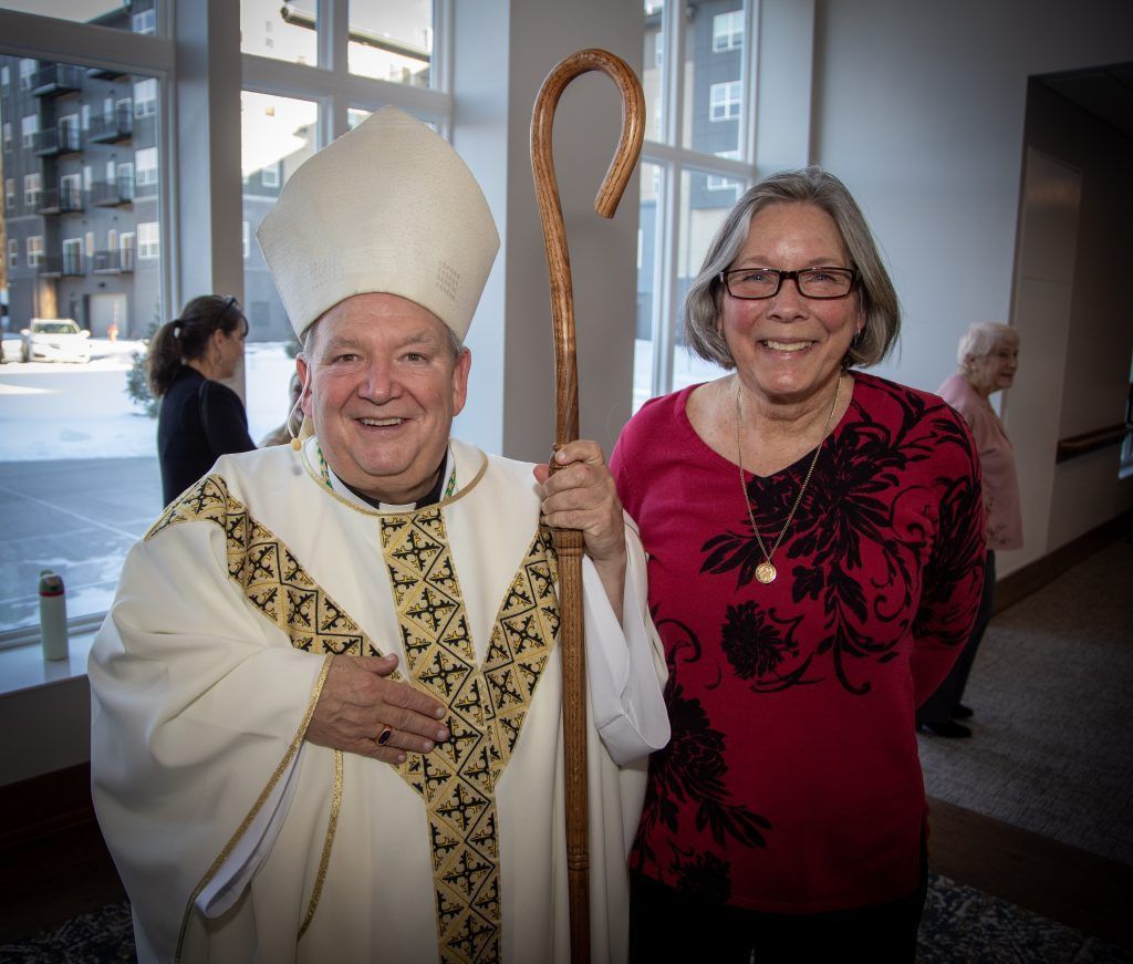 A smiling bishop in white liturgical vestments holding a wooden crozier stands beside a person in a red patterned top.