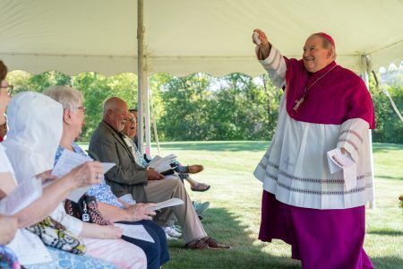 A bishop in fuchsia and white liturgical vestments holds up a white wafer while facing an audience seated under a tent.