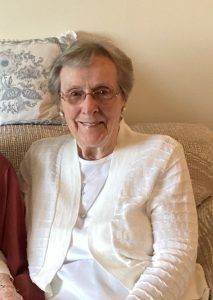 A person with light hair and glasses wearing a white cardigan and shirt, sitting on a patterned sofa.