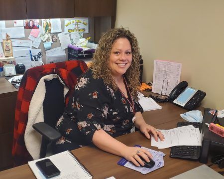 A smiling person with curly hair wears a floral top while sitting at a desk with a computer mouse and papers.
