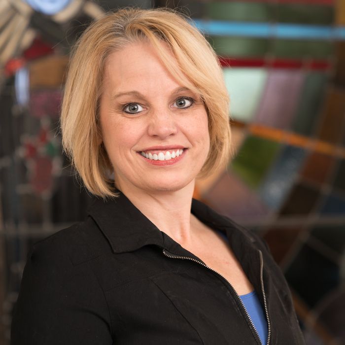 A smiling woman with blonde hair, wearing a black collared jacket over a blue top, posed against a stained-glass backdrop.