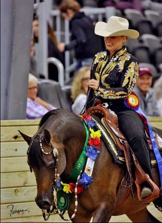 A rider in a formal western show outfit and hat rides a horse decorated with flower garlands and ribbons in an arena.