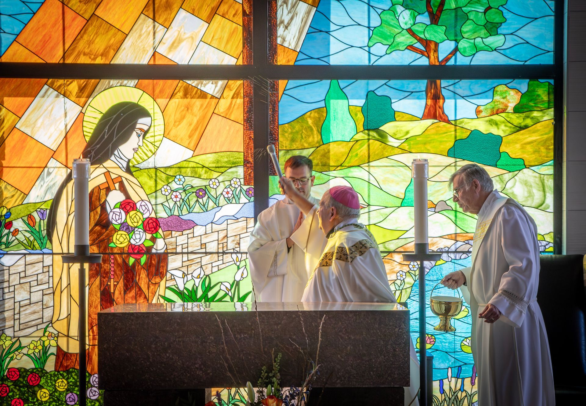 A bishop and clergy perform a ritual rite at a stone altar in front of a colorful stained-glass window.