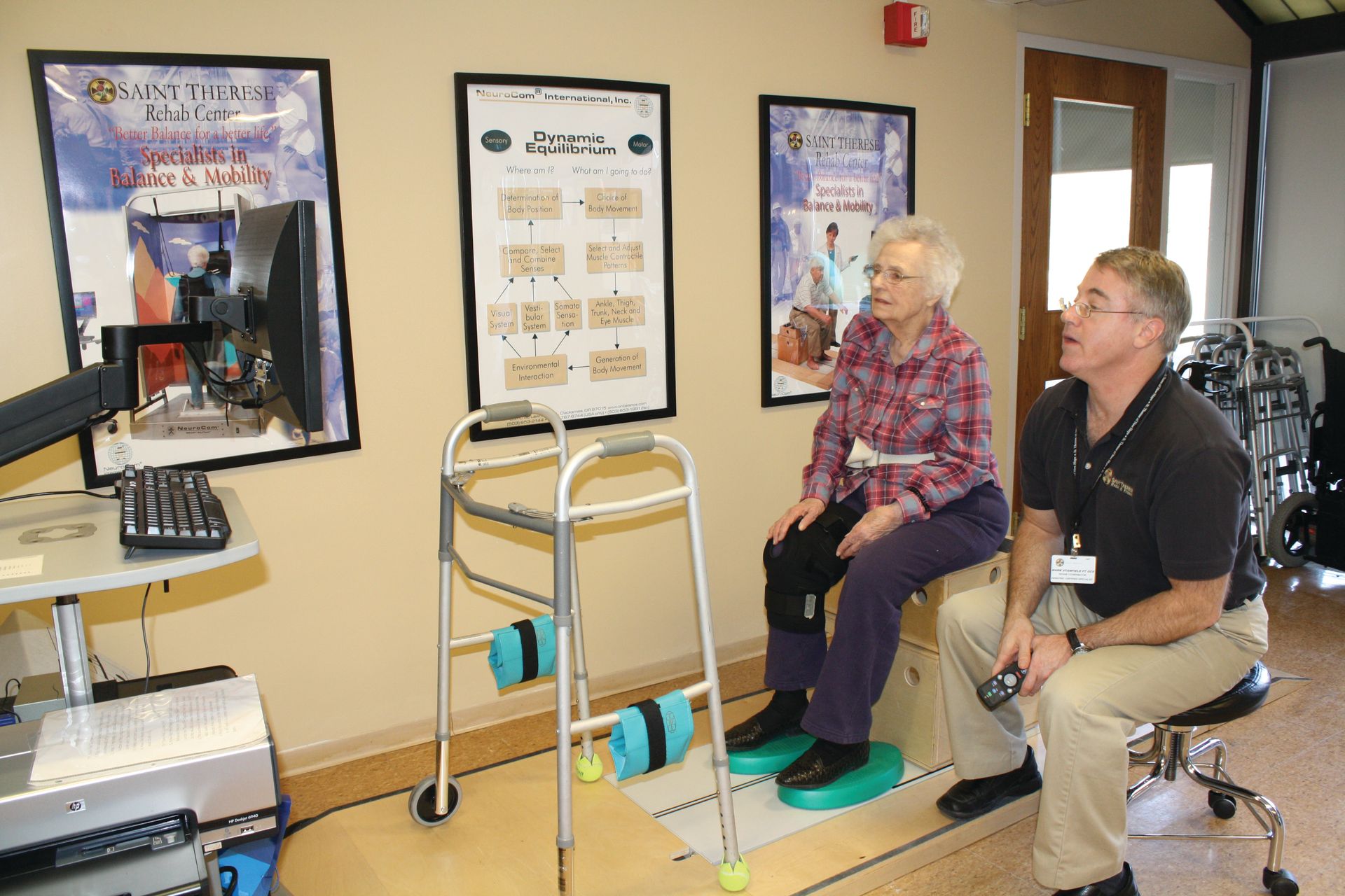 A professional in a clinical setting assists a person seated on a stool while using a walker and platform for therapy.