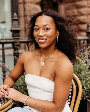 Smiling woman seated outdoors at a café, wearing a white strapless top and gold jewelry.