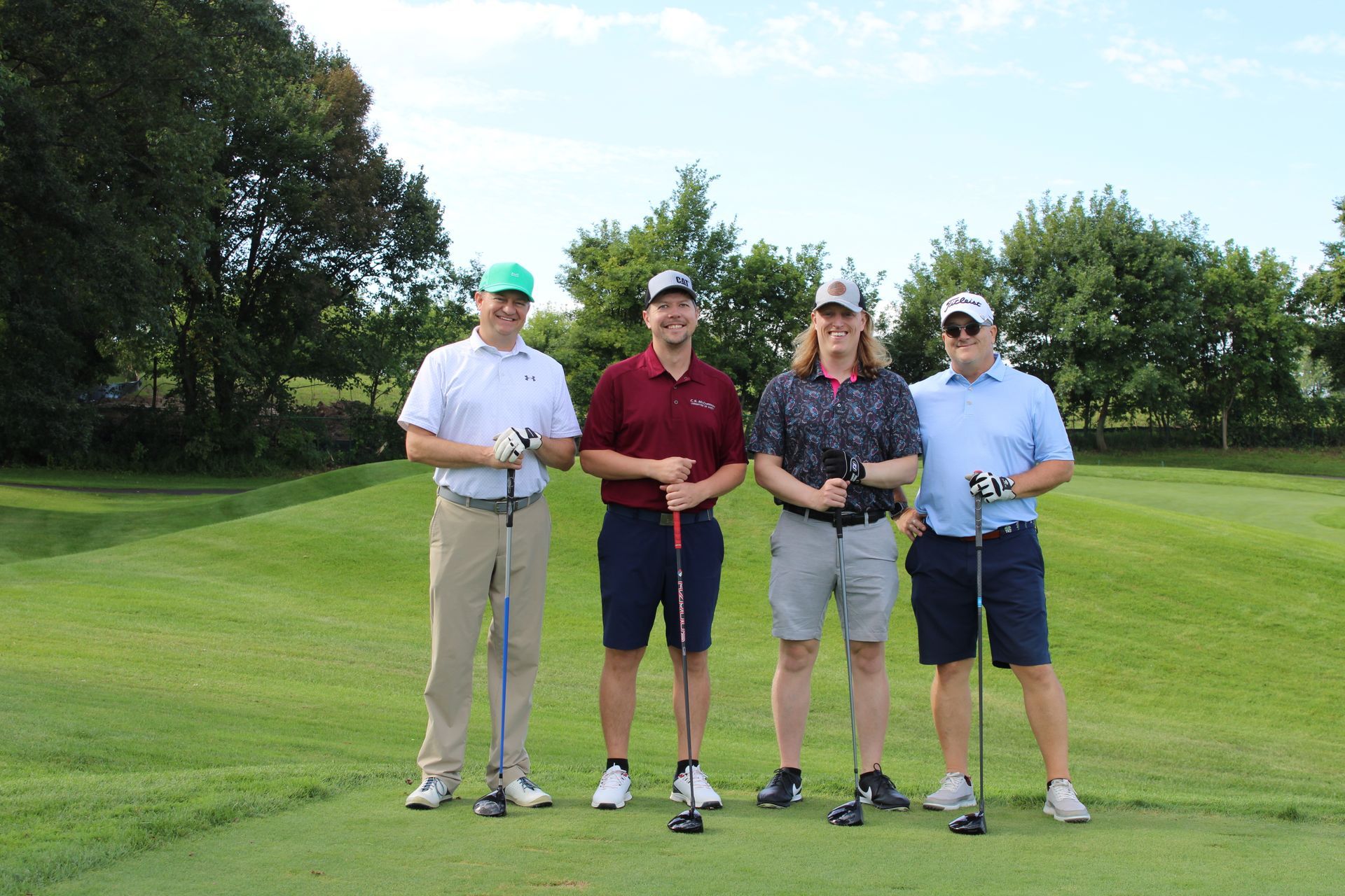 Four golfers stand on a grassy golf course, each holding a club and smiling toward the camera.