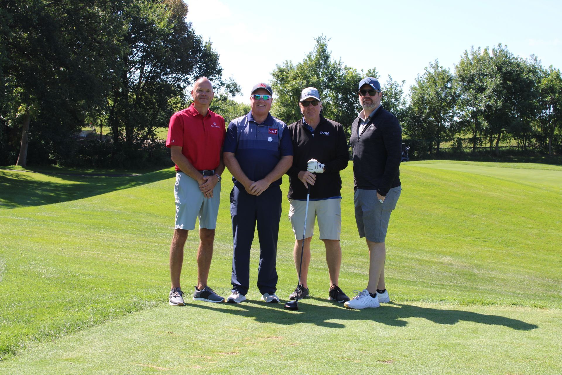Four people stand in a line on a sunny golf course, posing for a photo on the green.