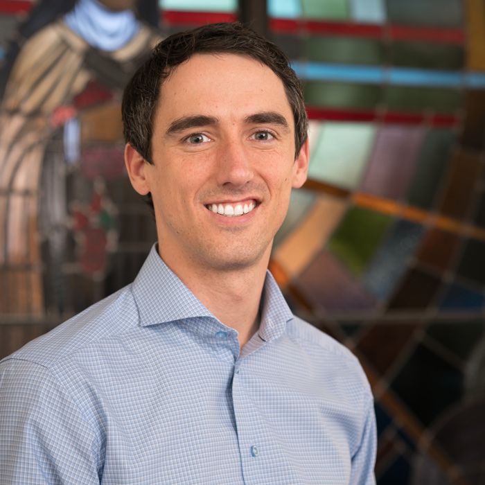 A person with short dark hair smiling in a light blue button-down shirt, set against a blurred stained glass background.
