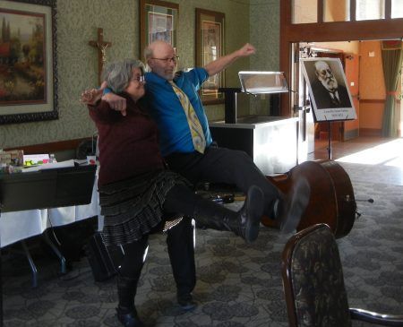 Two people perform a dance step with arms extended in a room featuring a portrait of Sigmund Freud and a double bass.