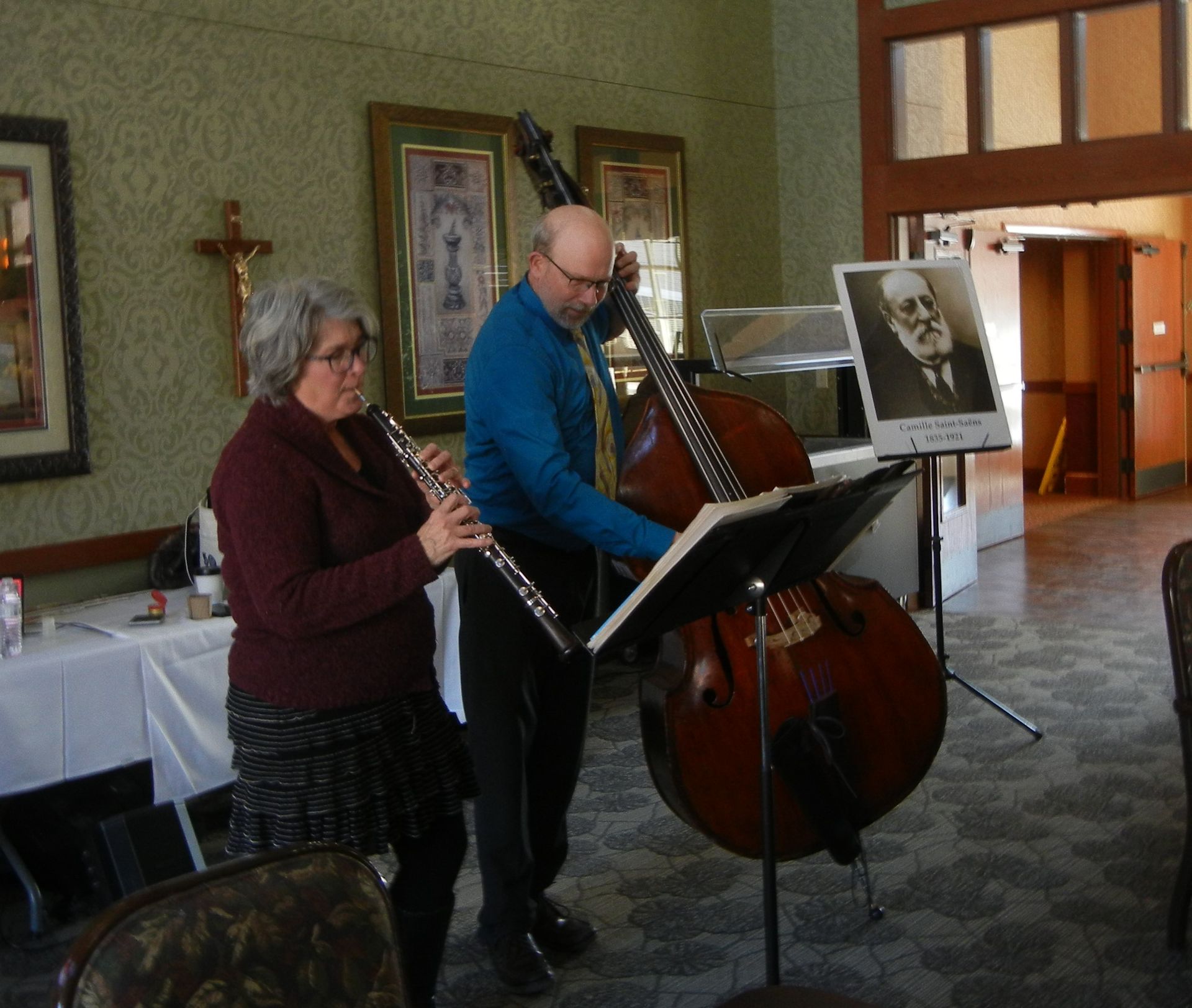 Two musicians perform together: one plays the flute while the other plays an upright bass, standing before music stands.