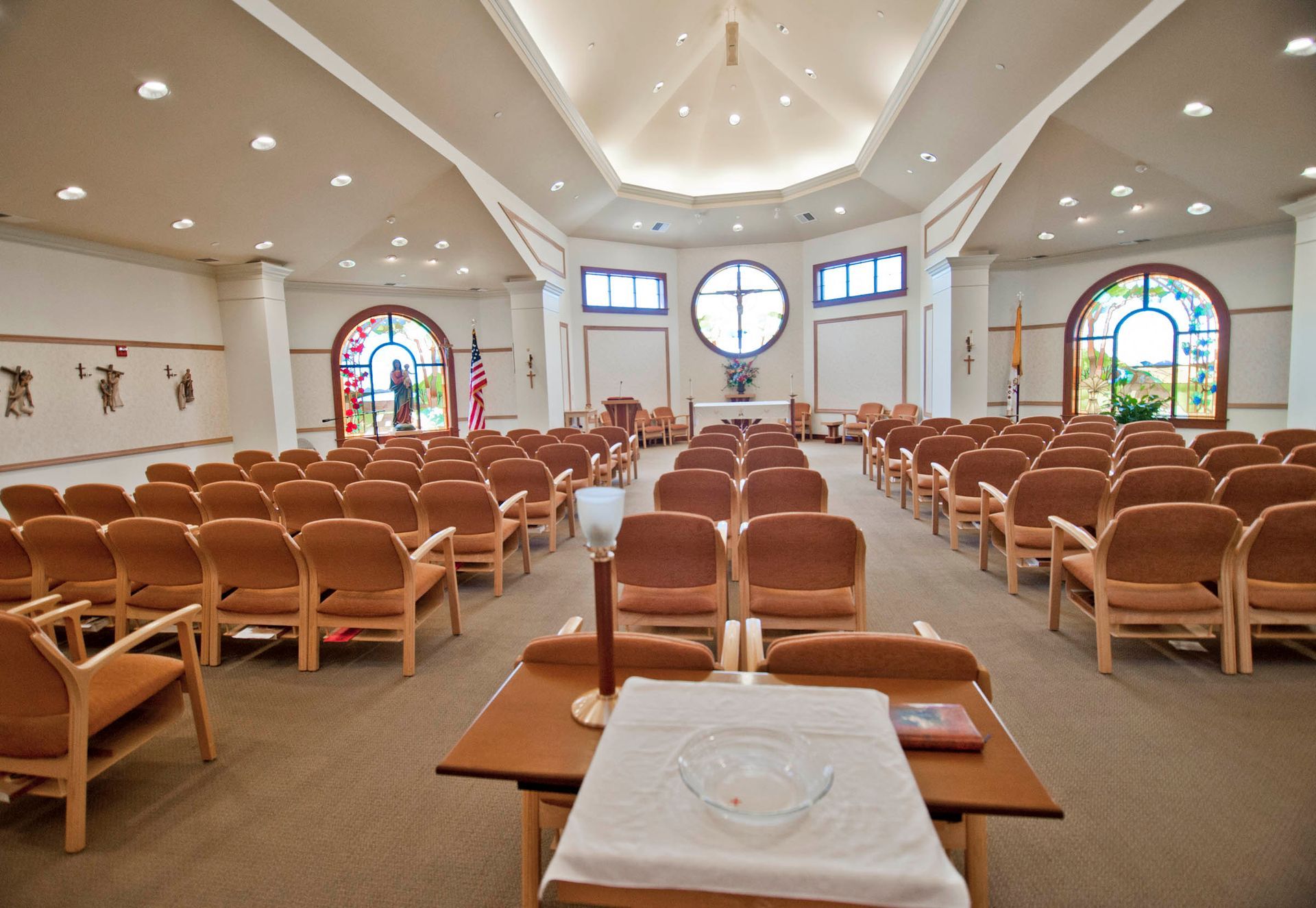 Rows of chairs face a central altar in a modern, brightly lit chapel with stained-glass windows.