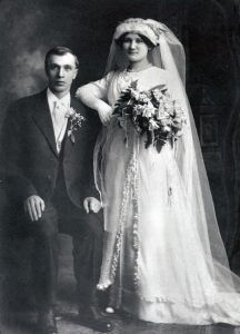 A black-and-white formal wedding portrait showing a seated groom and a standing bride holding a large bouquet.