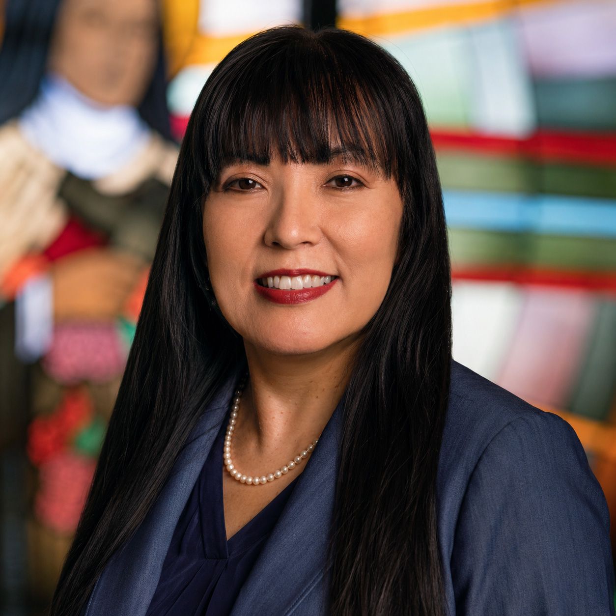 A smiling person wearing a blue blazer and pearl necklace, posed against a colorful, out-of-focus background.