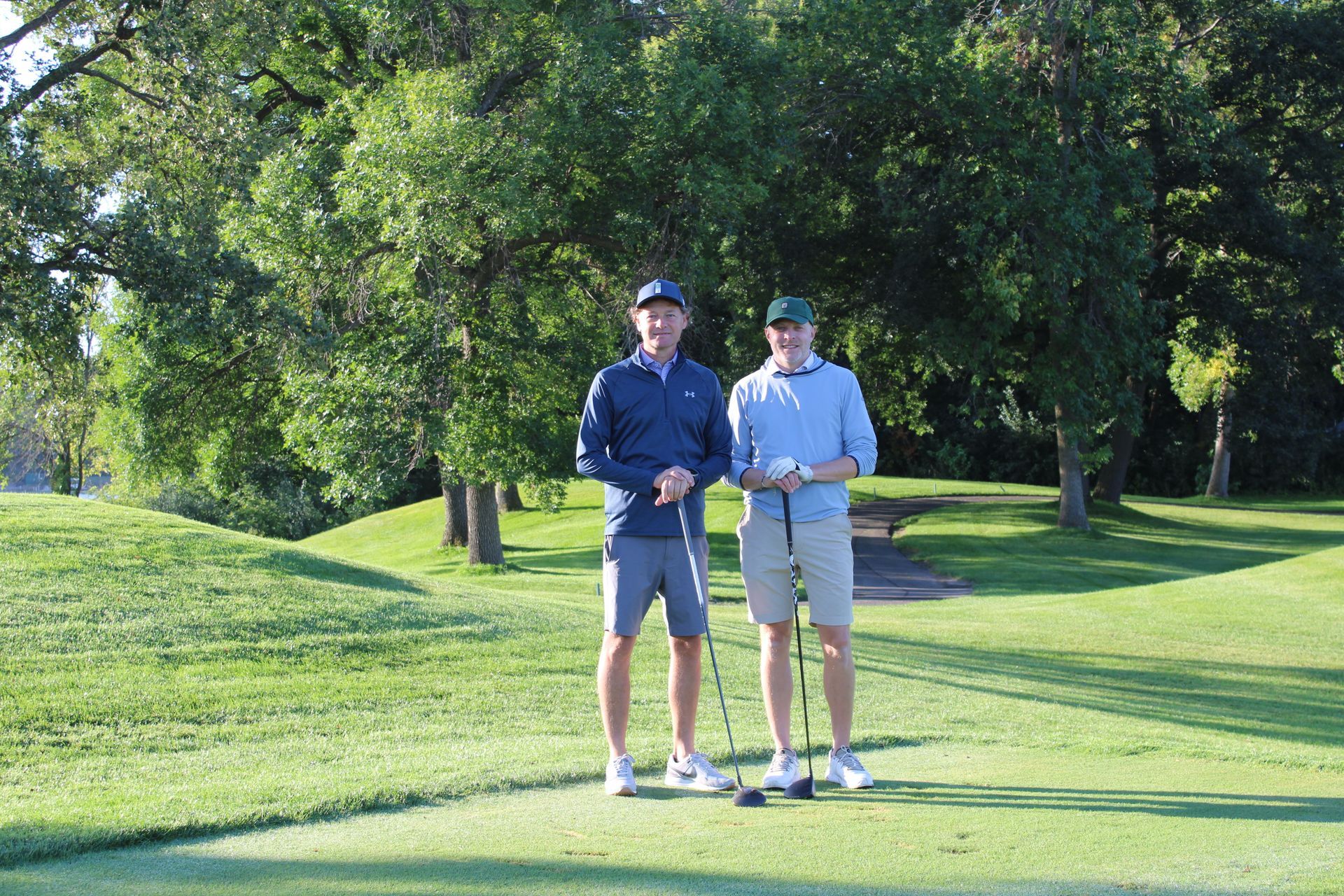 Two people in golf attire standing on a sunny golf course holding their clubs.