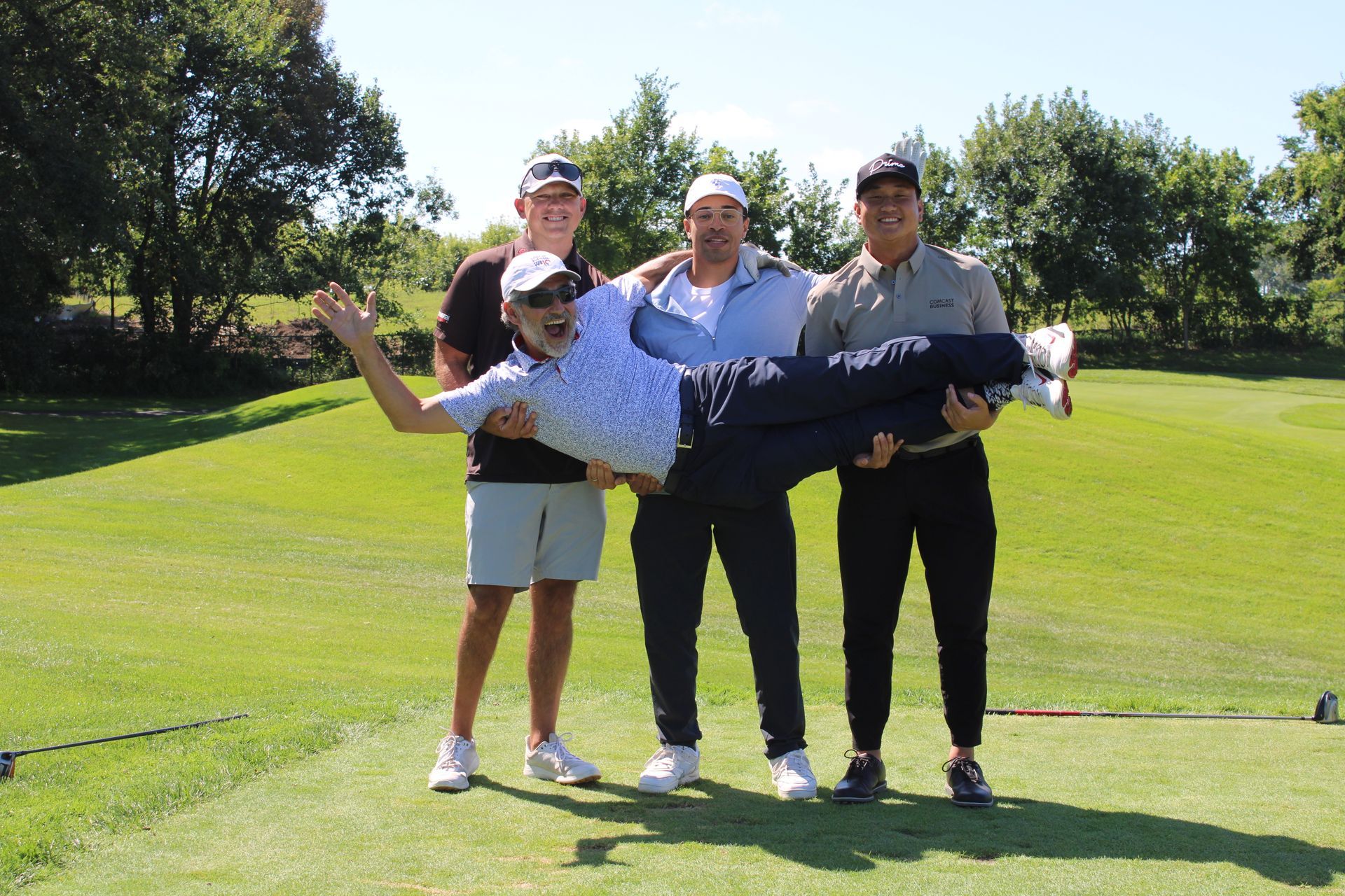 Three people hold a fourth person horizontally in their arms while standing on a sunny golf course.