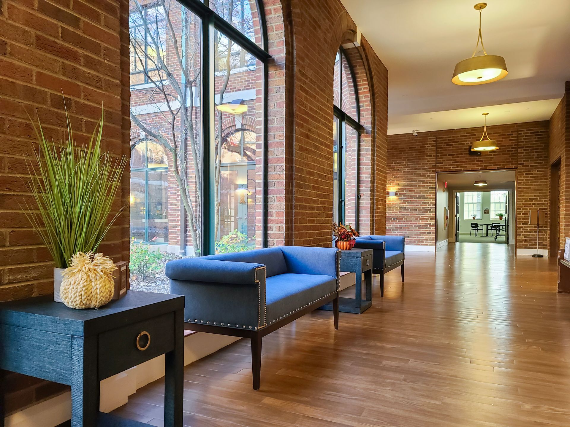 Hallway with brick walls and large windows, featuring blue seating, decorative plants, and pendant lighting.